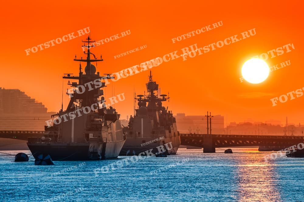 На фотографии изображены военные корабли на фоне заката в Санкт-Петербурге. Солнце опускается за горизонт, окрашивая небо в яркие оранжевые оттенки, создавая драматический и впечатляющий вид. Фото идеально подходит для использования в туристических материалах, статьях о Санкт-Петербурге и военной тематике.